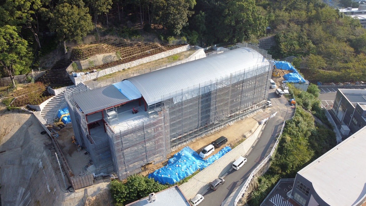 【上空から見た、新社屋の今】 緑に囲まれた敷地に、アーチ屋根のシルエットがくっきり🚁 足場の内側では、カーテンウォールの骨組みづくりが進んでいます🏗️ 新社屋の“顔”となる外観が、少しずつ形になってきました✨